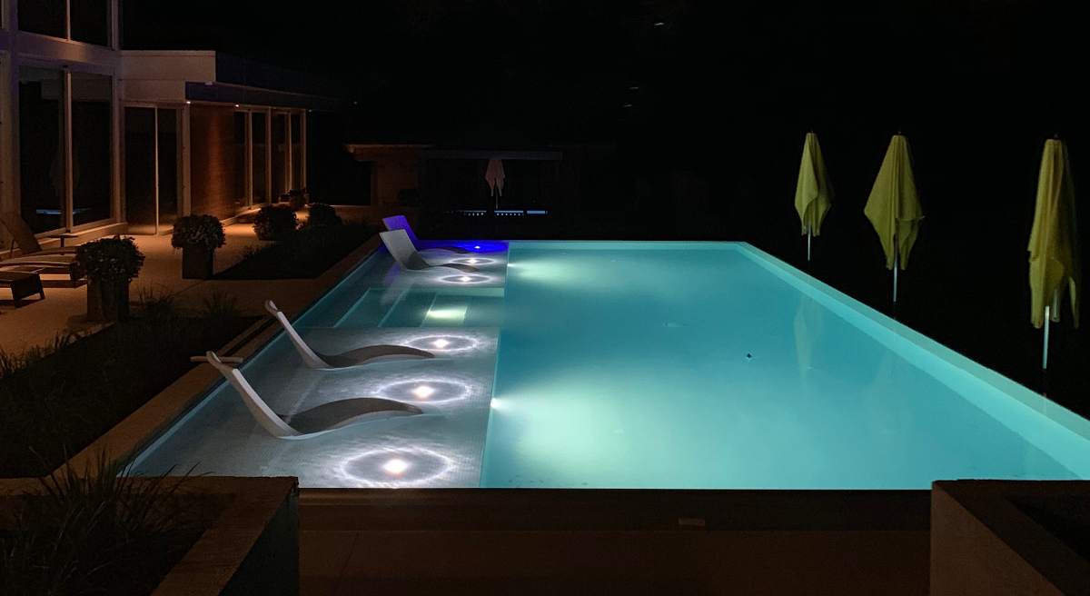 Night time view of Private Residence's backyard infinity pool with underwater lights in the pool and along the tanning ledge 